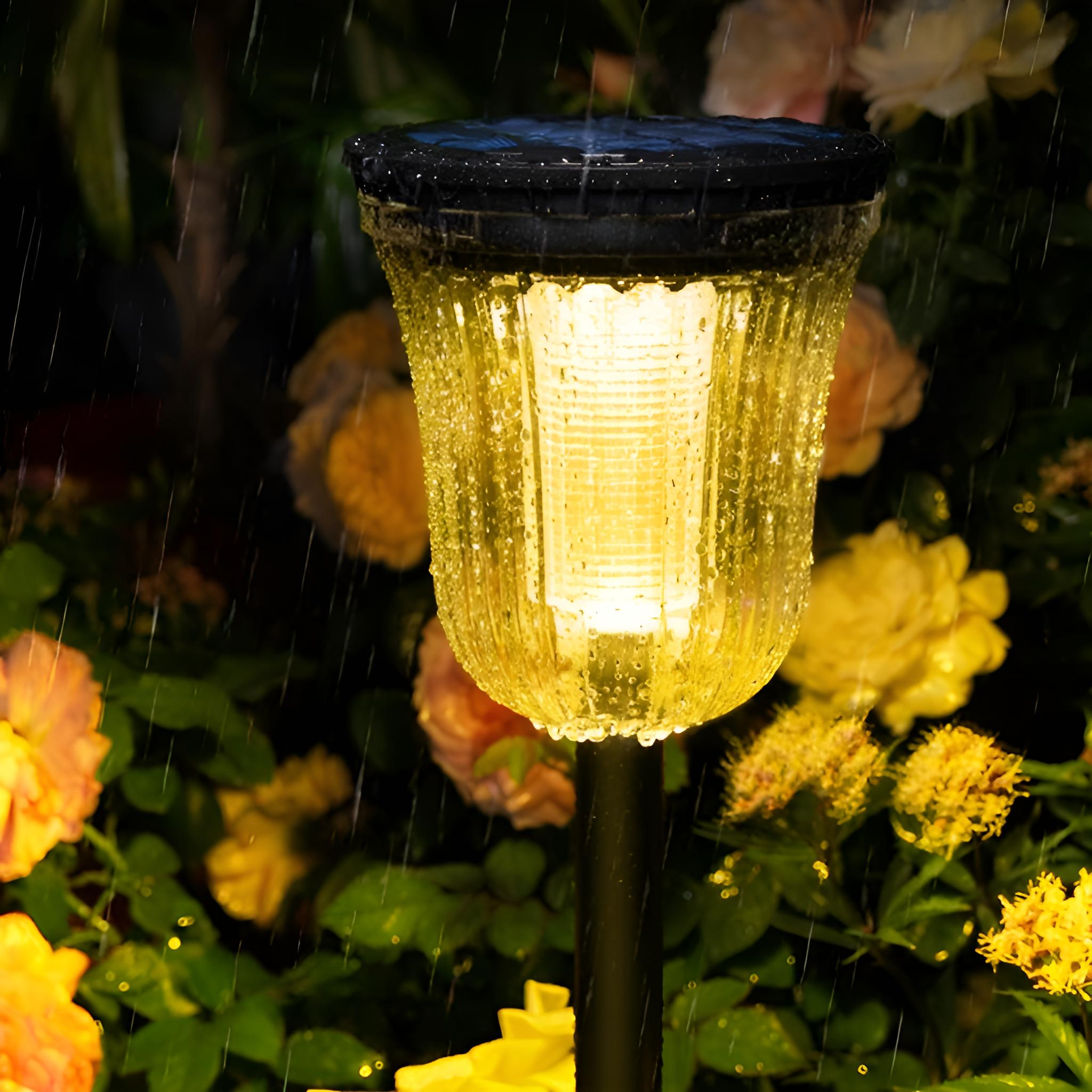 Eine Solar-Gartenleuchte bei Nacht. Sie ist beleuchtet und von Blumen umgeben, Regentropfen sind sichtbar.