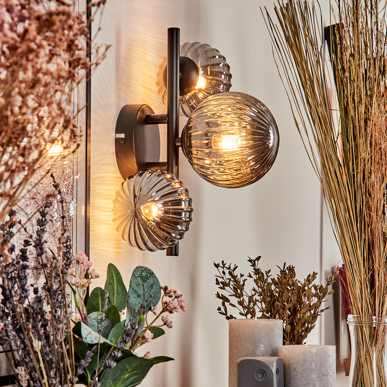 Eine Wandlampe mit drei Glaskugeln. Sie befindet sich neben Blumen und Pflanzen.