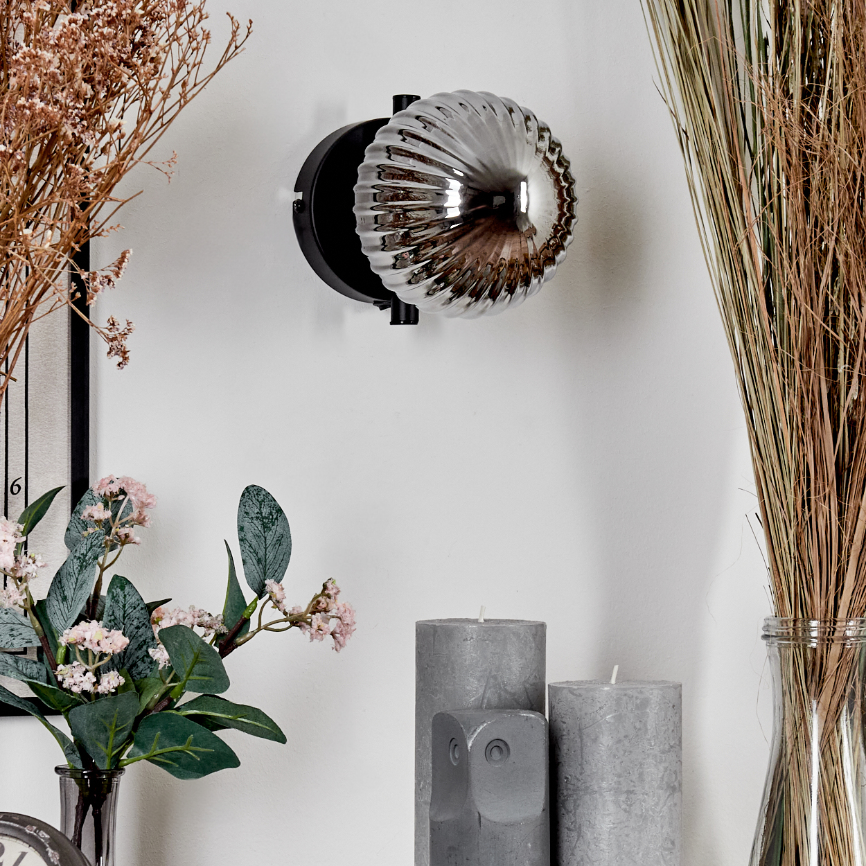 Eine Wandleuchte mit Glaskugel. Die Lampe ist an einer weißen Wand montiert, mit Kerzen und Blumen im Vordergrund.