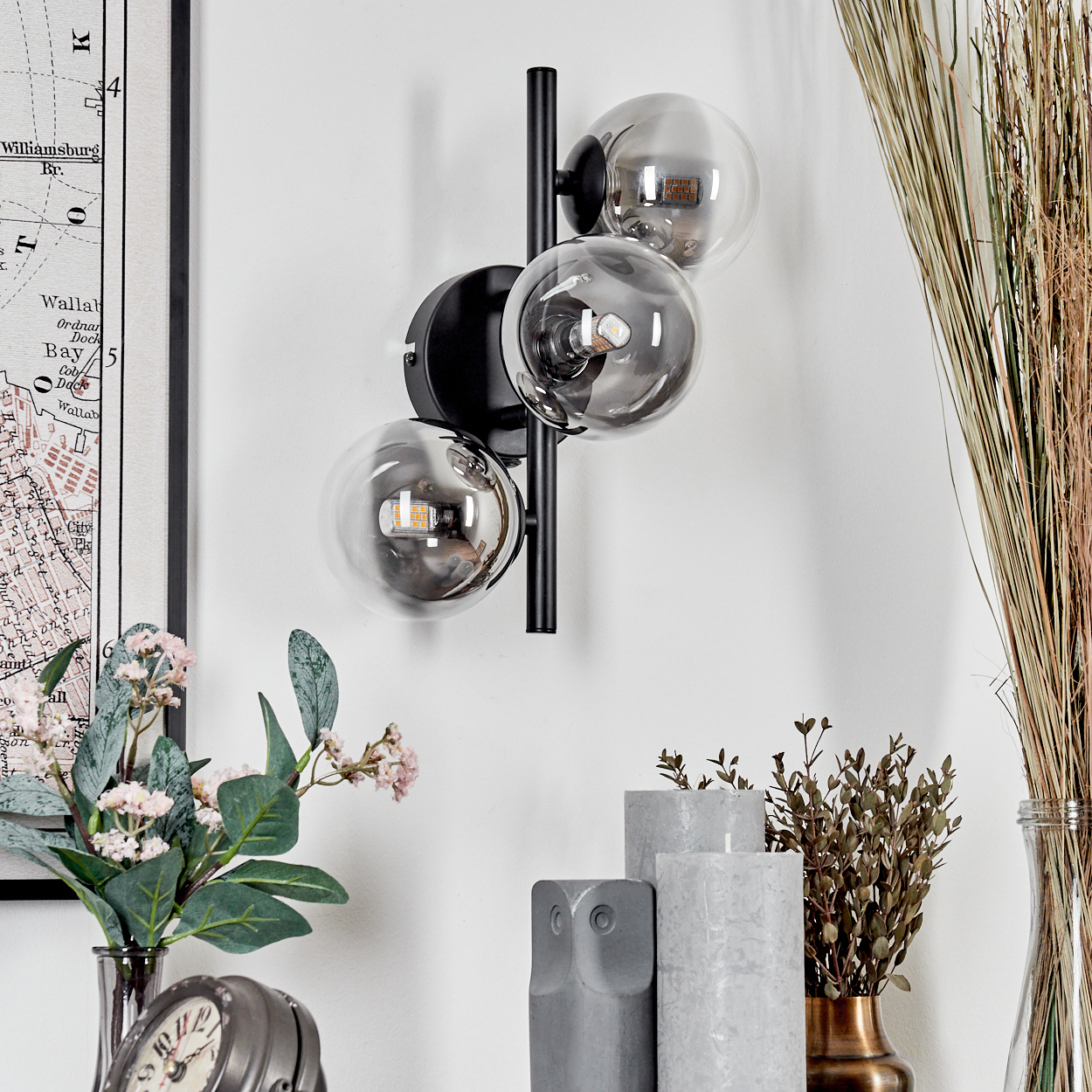 Schwarze Wandlampe mit drei transparenten, kugelförmigen Glühbirnen. Deko mit Blumen, Karte, Kerzen.