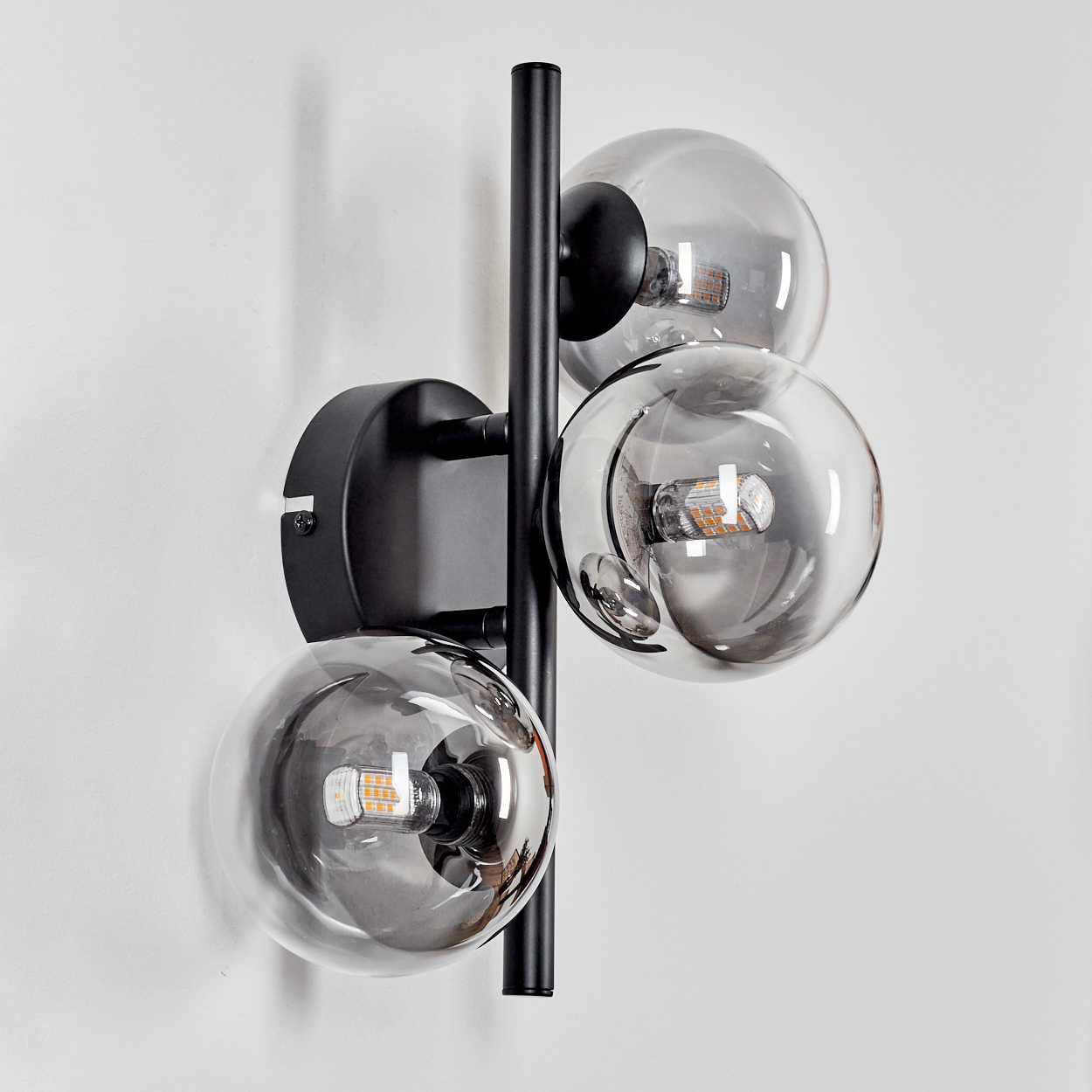 Eine schwarze Wandlampe mit drei klaren, kugelförmigen Glühbirnen. Die Lampe ist an einer weißen Wand montiert.