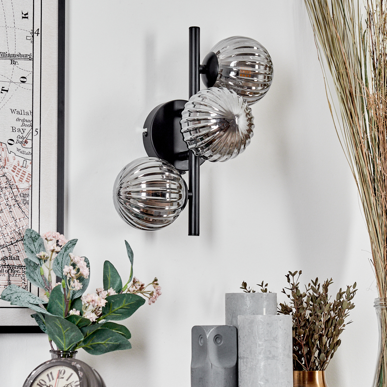 Wandlampe mit drei Kugelleuchten. Trockenpflanzen, Kerzen und eine Uhr sind sichtbar.