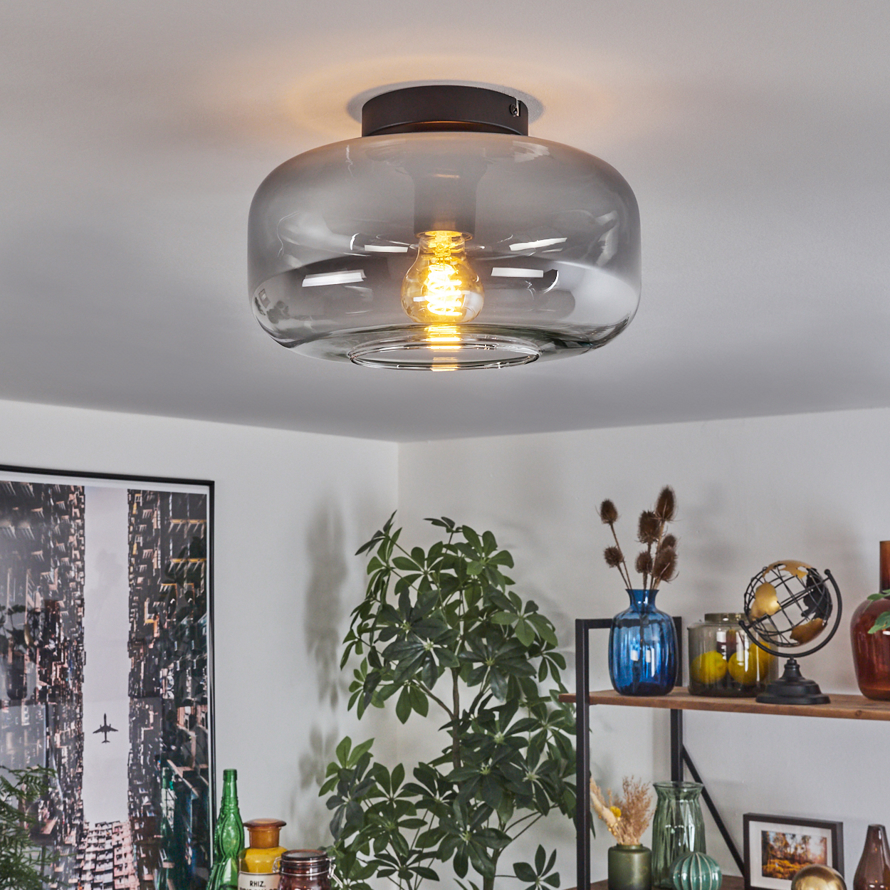 Deckenleuchte mit klarem Glasschirm, leuchtender Glühbirne und schwarzem Sockel. Vor einer weißen Decke.