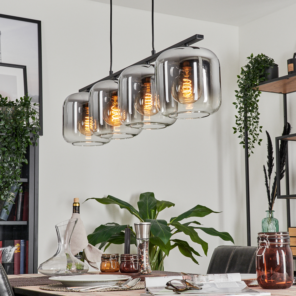 Vier Lampen mit rechteckigen Glasschirmen, die über einem Esstisch mit Dekoration hängen.