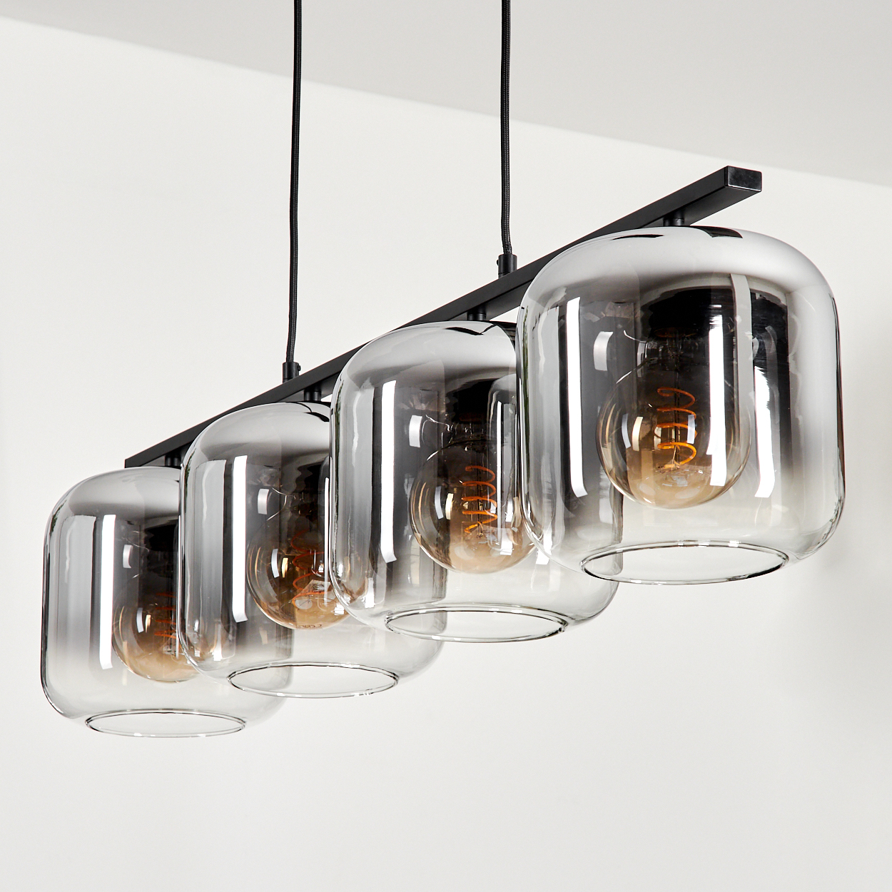 Vier Lampen mit rechteckigen Glasschirmen an einer schwarzen Stange, jede mit einer sichtbaren Wendelglühbirne.