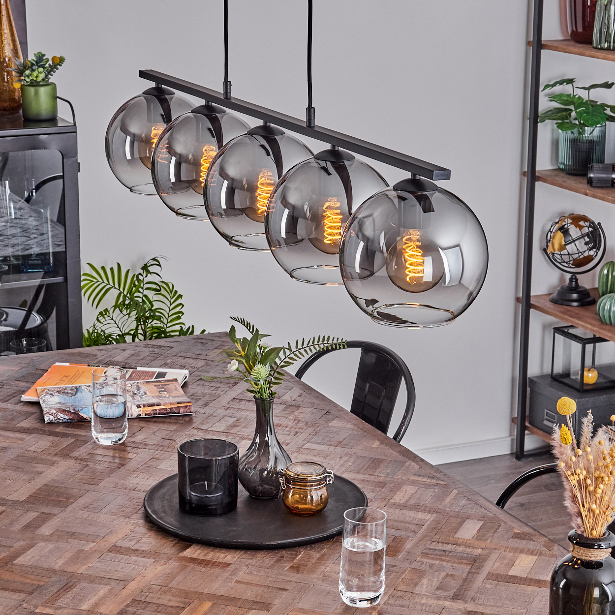 Ein Esszimmer mit einem dunklen Holztisch und fünf Pendelleuchten mit grauen Glaskugeln. Gläser und Dekorationen sind aufgestellt.