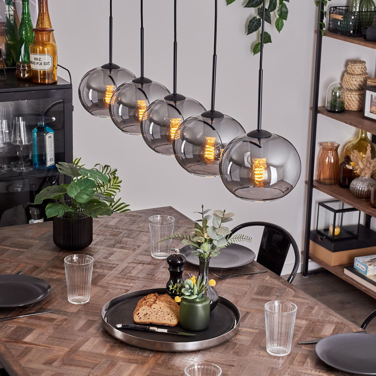 Esszimmer mit Holztisch, Glaswaren und Pflanzen. Fünf Hängelampen mit grauen Glasschirmen.