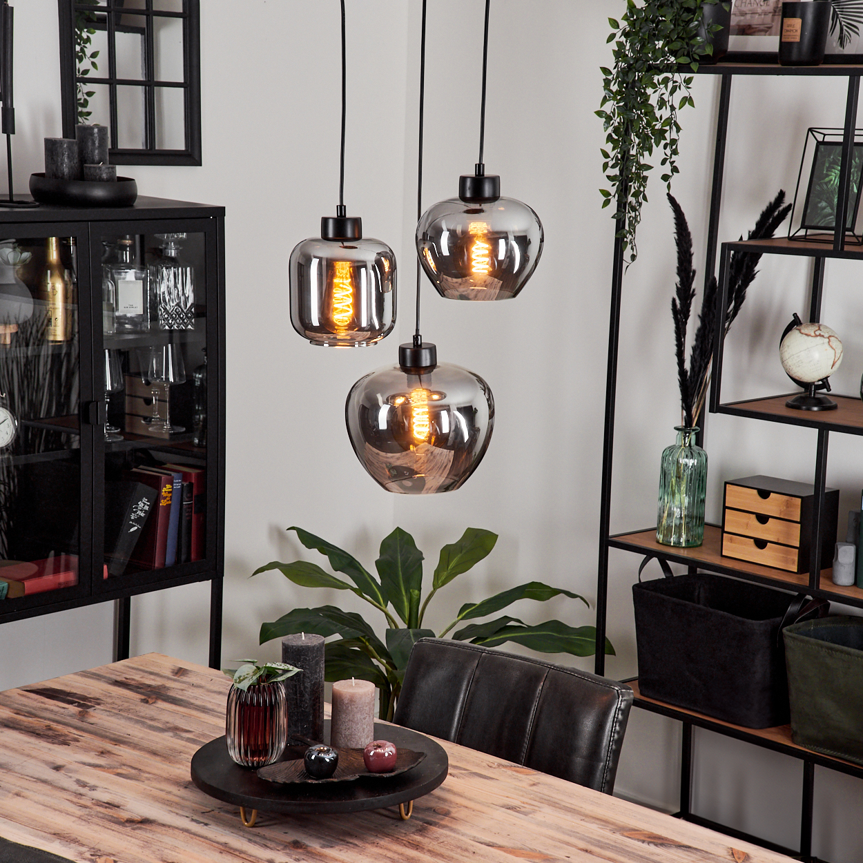 Drei Pendelleuchten mit Holztisch und Dekoration. Jede Leuchte hat eine schwarze Halterung und einen klaren, bauchigen Glasschirm.