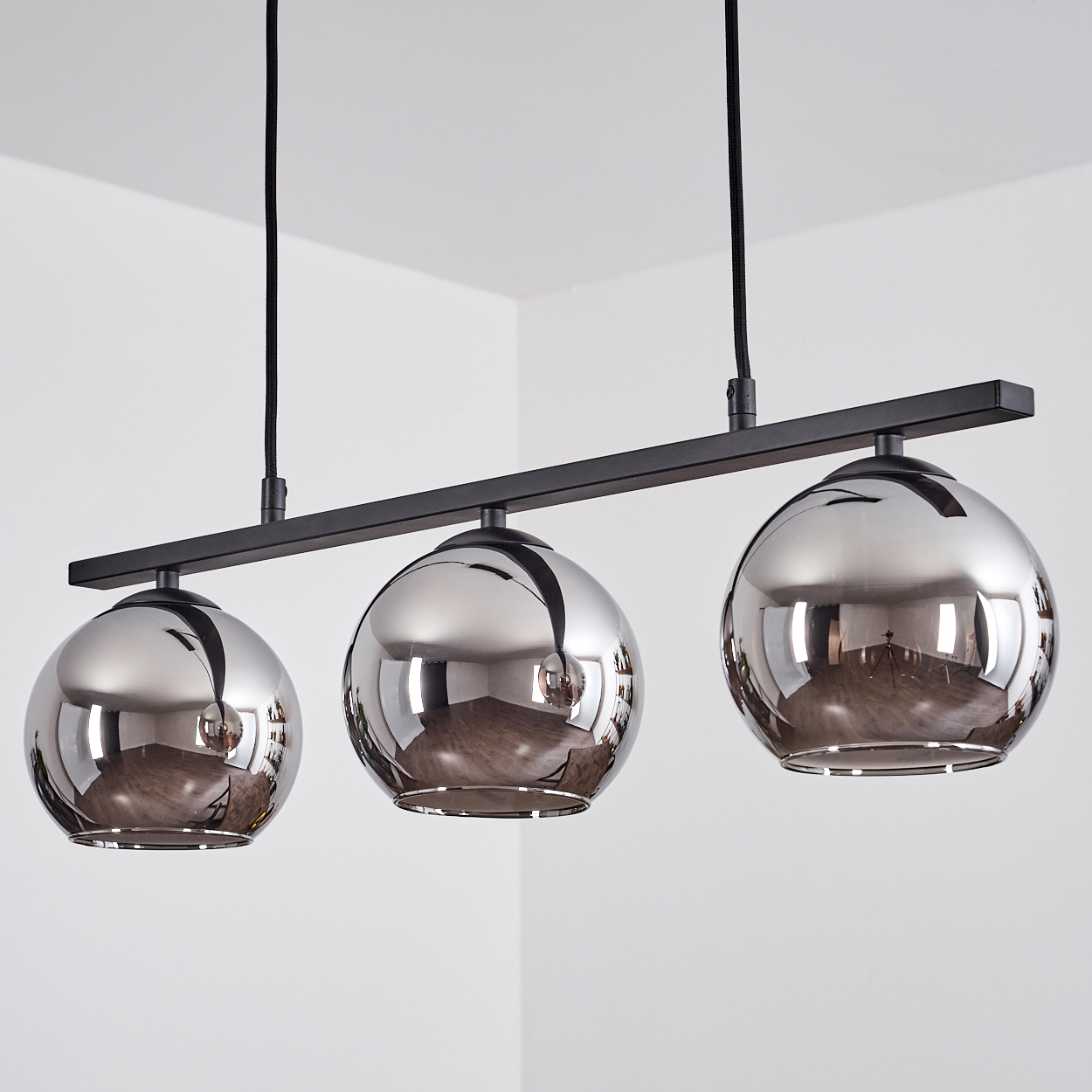 Drei silberne Kugellampen hängen an der Decke. Schwarze Stangen und Kabel. Weißer Wandhintergrund.