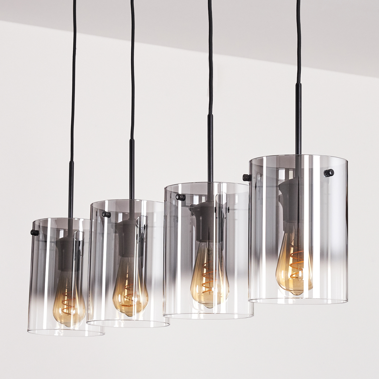 Vier zylindrische Pendelleuchten mit freiliegenden Glühbirnen hängen von einer Decke. Die Lichter sind an.