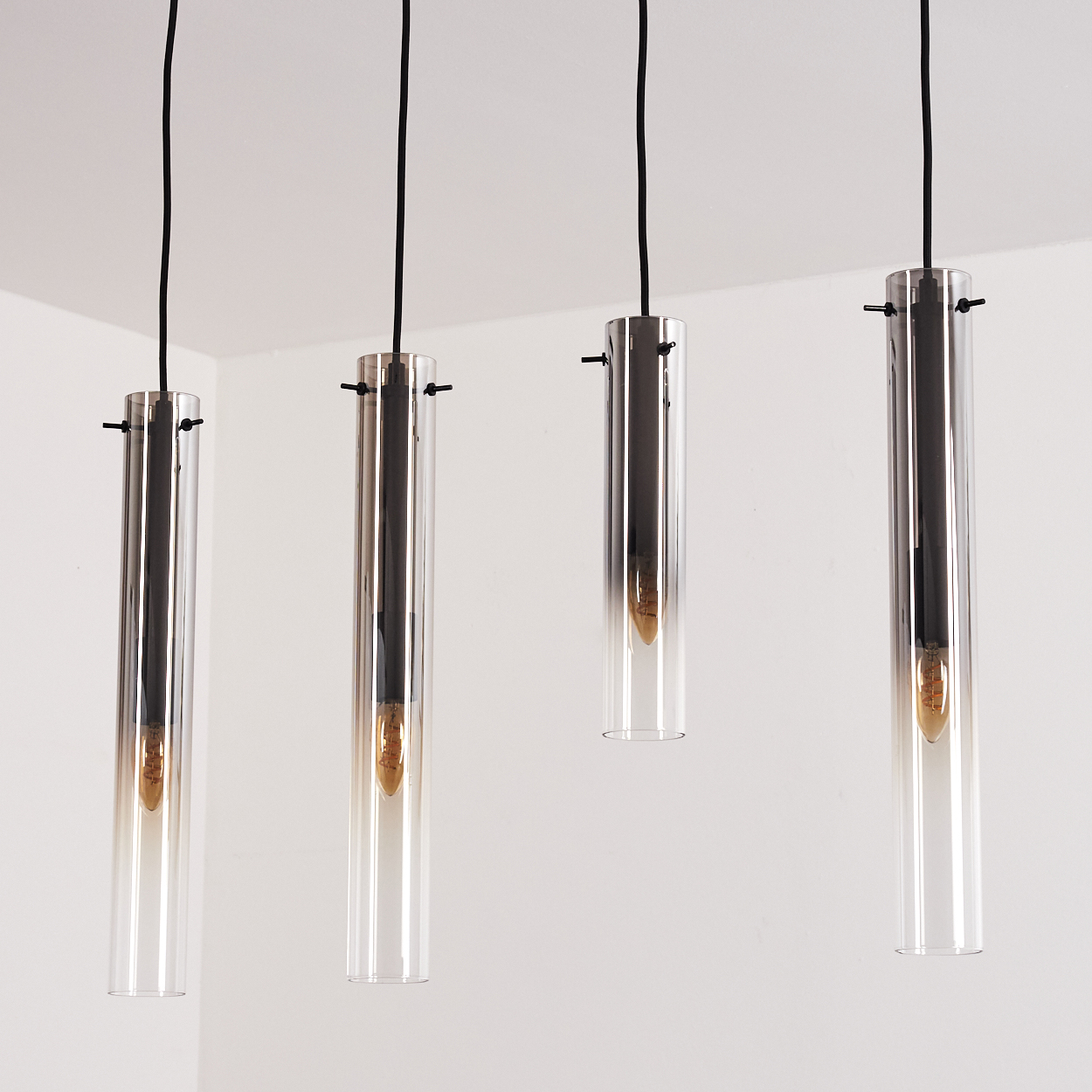 Vier Pendelleuchten, die von einer weißen Decke hängen. Die Leuchten haben klare, zylindrische Glasschirme und bernsteinfarbene Glühbirnen.