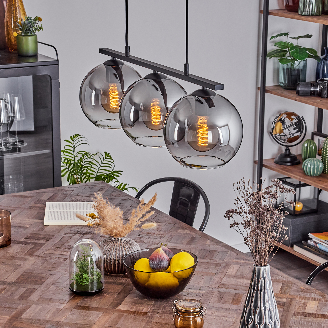 Drei verchromte Kugelhängeleuchten hängen über einem Holztisch mit Deko und Pflanzen. Innenraum.