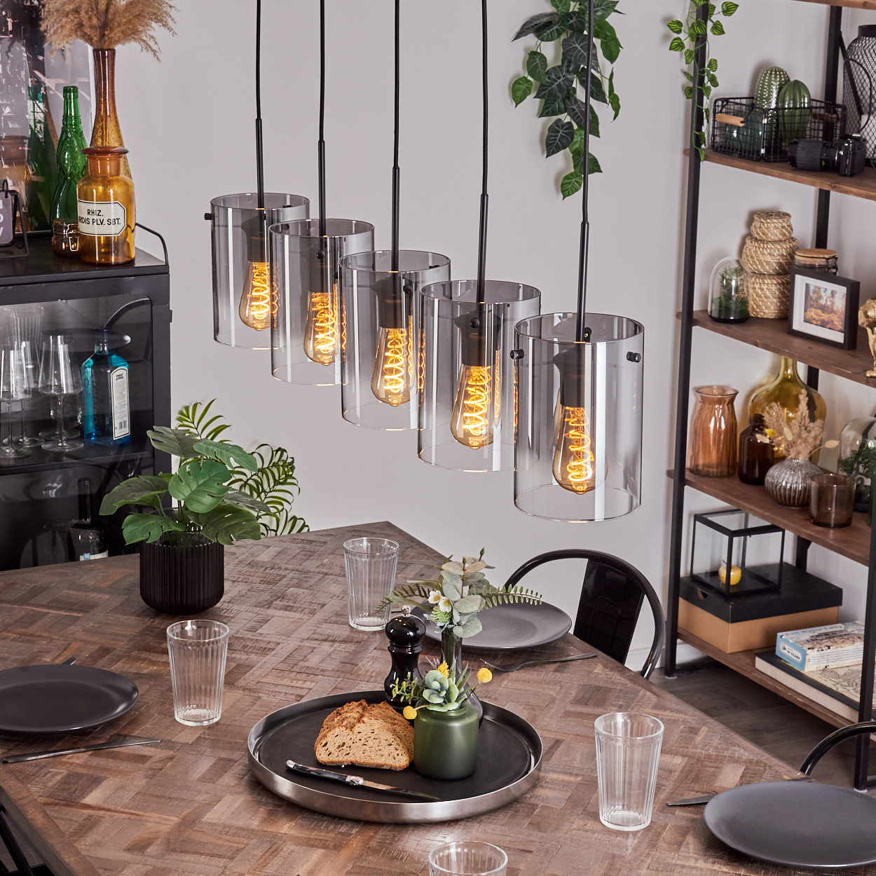 Fünf Pendelleuchten mit zylindrischen, klaren Glasschirmen hängen über einem Holztisch mit Tellern und Essen.