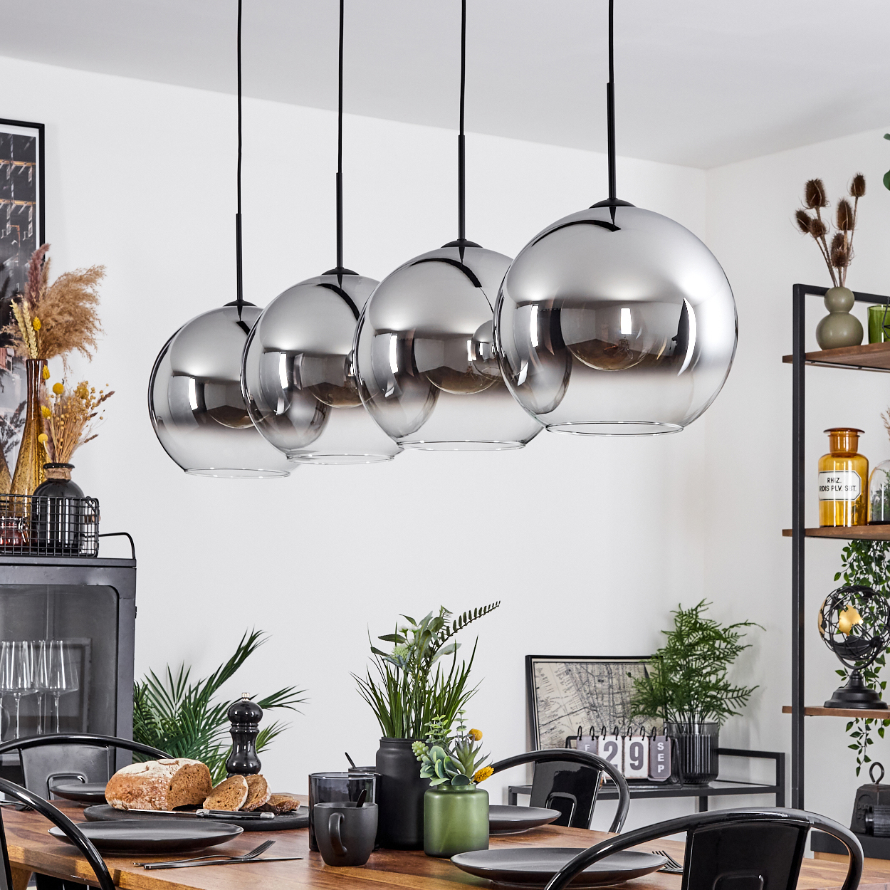 Vier Pendelleuchten mit kugelförmigen, verspiegelten Glasschirmen hängen über einem Holztisch mit Tellern und einer Pflanze.