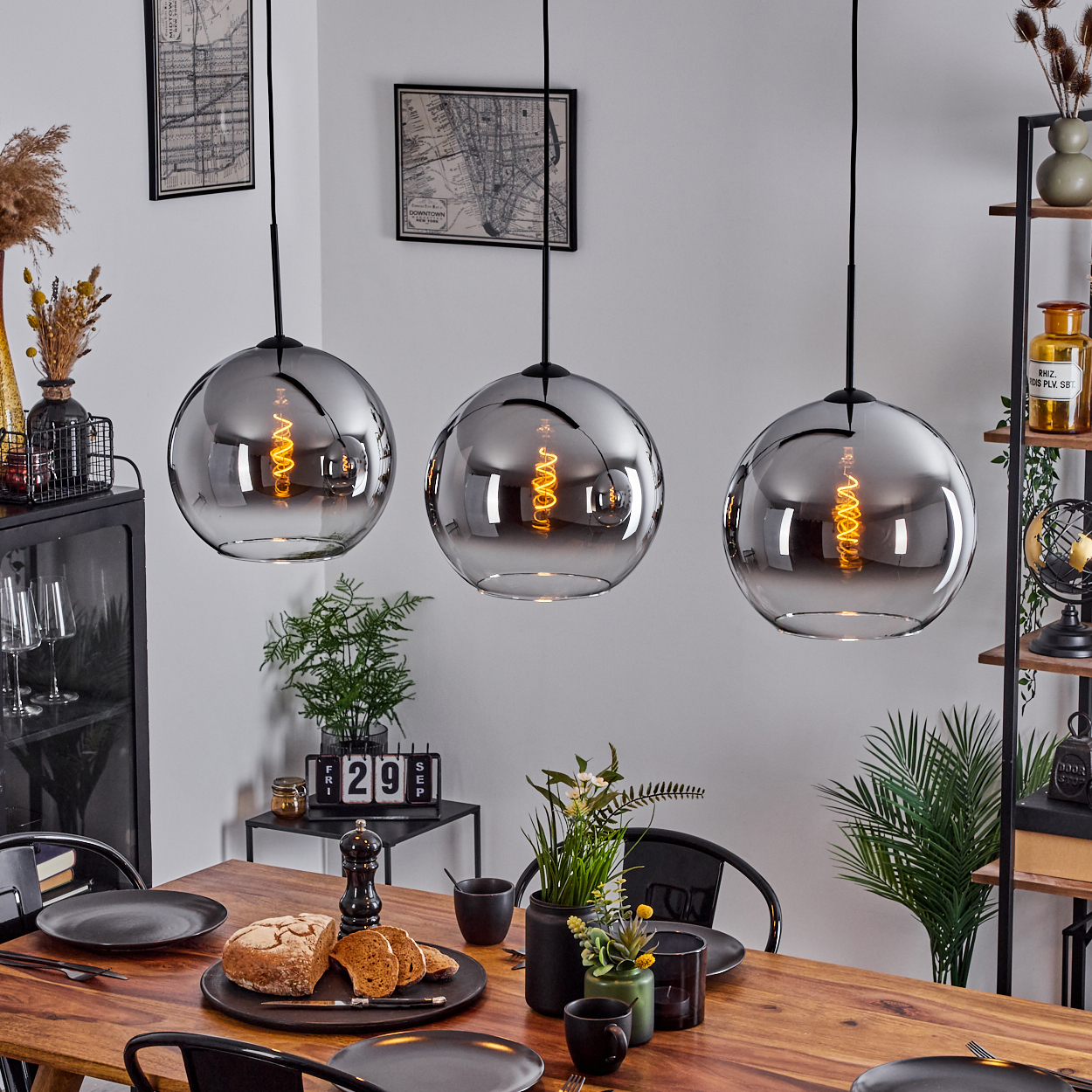 Drei silberne Pendelleuchten mit sichtbaren Glühbirnen hängen über einem Esstisch mit Essen, Blumen und Dekoration.