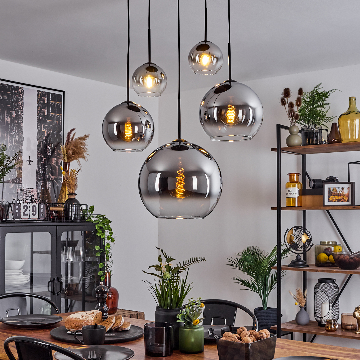 Fünf silberne Pendelleuchten mit sichtbaren Glühbirnen hängen über einem Esstisch mit Essen, Blumen und Dekoration.