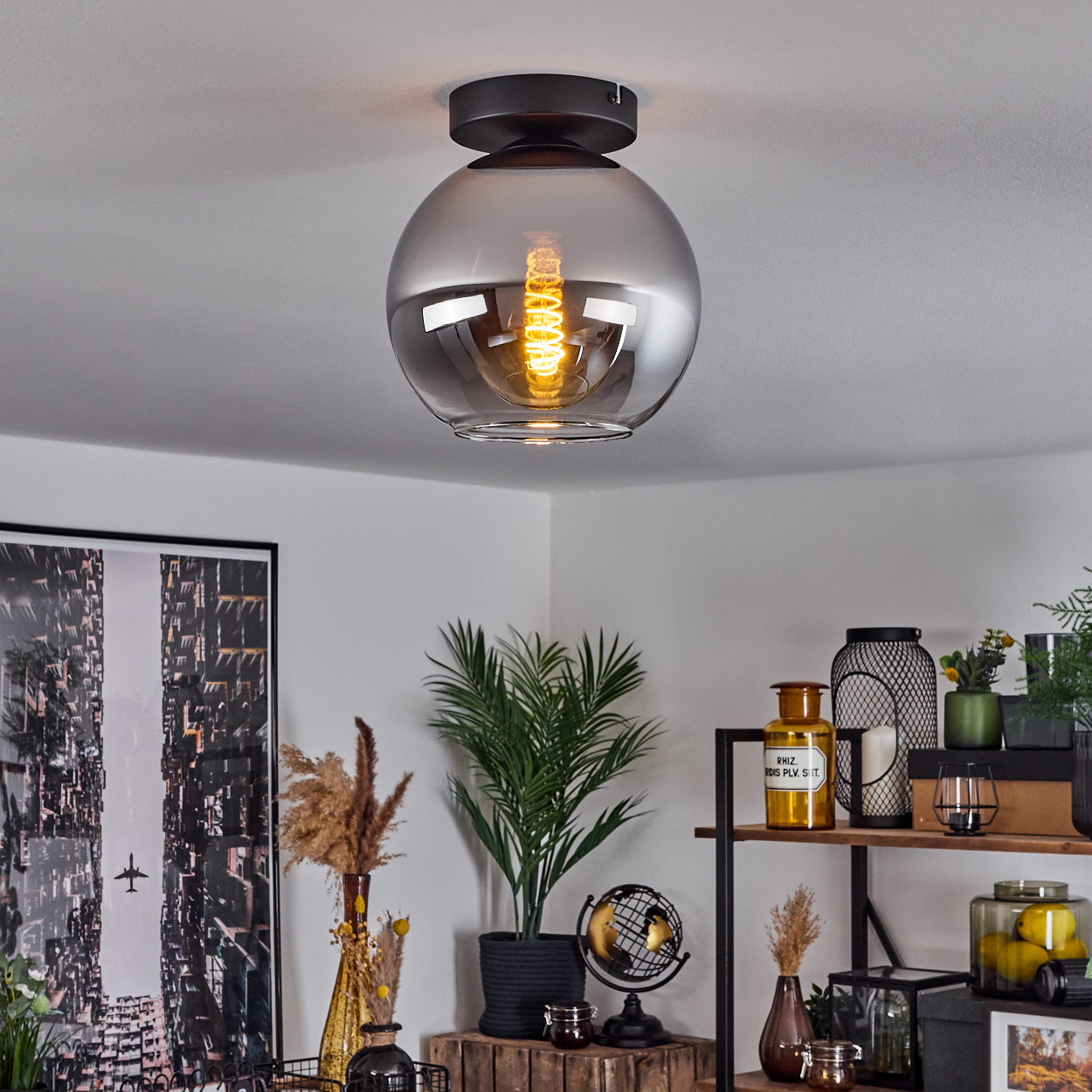 Eine Lampe, die von einer weißen Decke hängt, mit Dekoration im Hintergrund. Die Lampe ist kugelförmig mit sichtbarer Glühbirne.