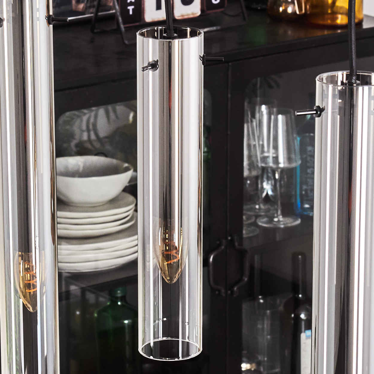 Drei zylindrische Glaslampen hängen und reflektieren den Raum. Ein schwarzer Schrank, Teller und eine Schüssel sind im Hintergrund.