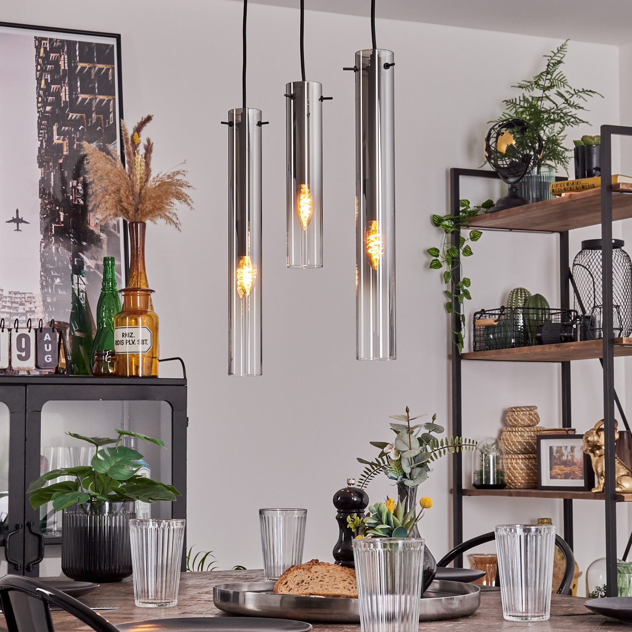 Drei Pendelleuchten mit Glasröhren über einem Holztisch. Esszimmer-Setting mit dekorativen Elementen.