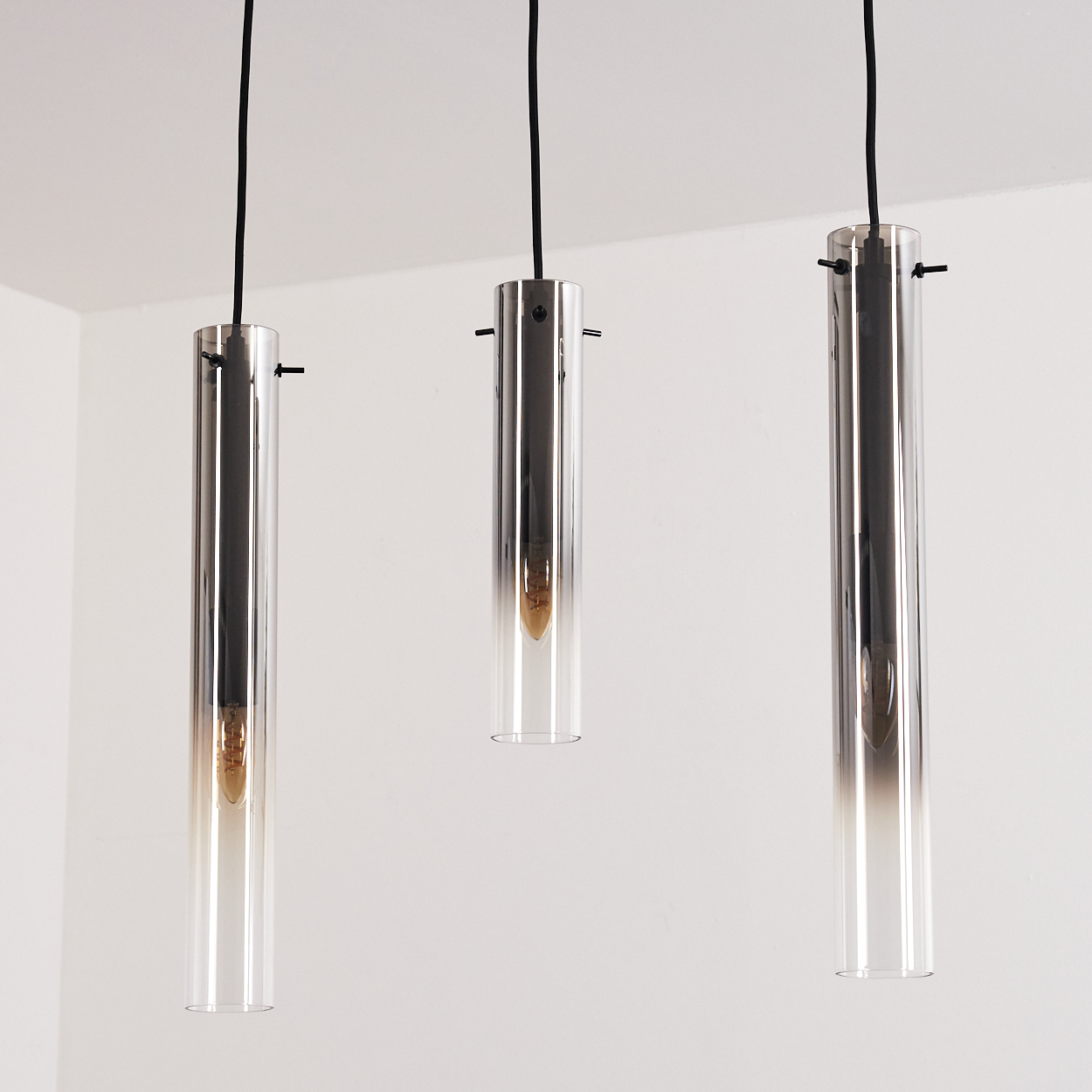 Drei transparente, zylindrische Pendelleuchten hängen vor einer weißen Wand, mit einer Glühbirne in jeder.