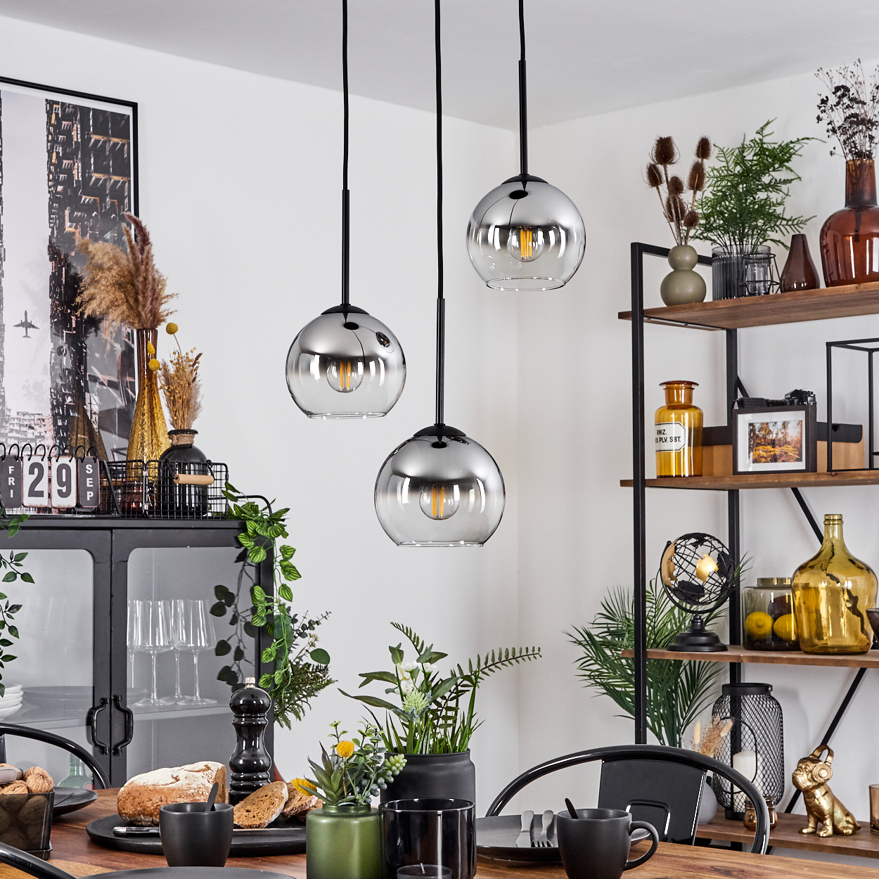 Ein Esszimmer mit Tisch, drei Pendelleuchten und Dekorationen. Ein Schrank und Regale sind sichtbar.