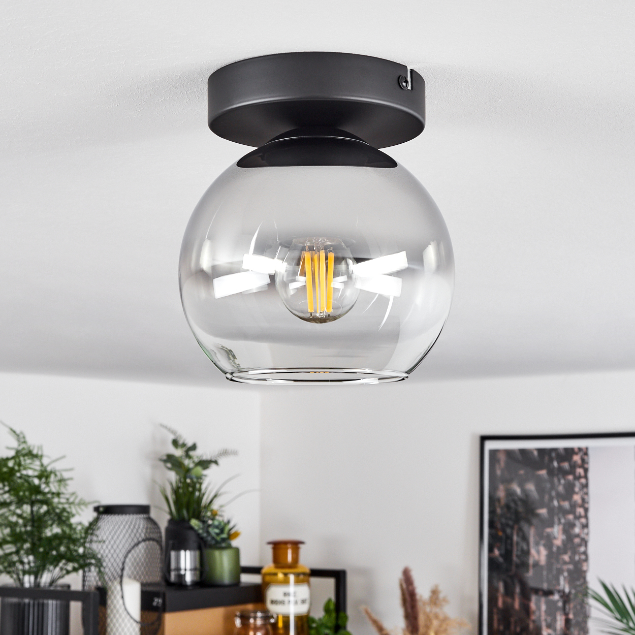 Eine Deckenleuchte mit einem Glasschirm und einer sichtbaren Glühbirne ist an einer weißen Decke montiert, umgeben von Dekoration.