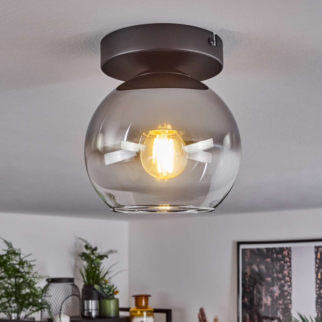 Eine Deckenleuchte mit einem Glasschirm und einer sichtbaren Glühbirne ist an einer weißen Decke montiert, umgeben von Dekoration.