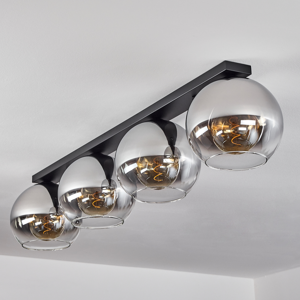Vier moderne Deckenleuchten. Jede Leuchte hat einen Glaskolben mit einer Glühbirne. Auf einem schwarzen Metallrahmen an einer weißen Decke montiert.