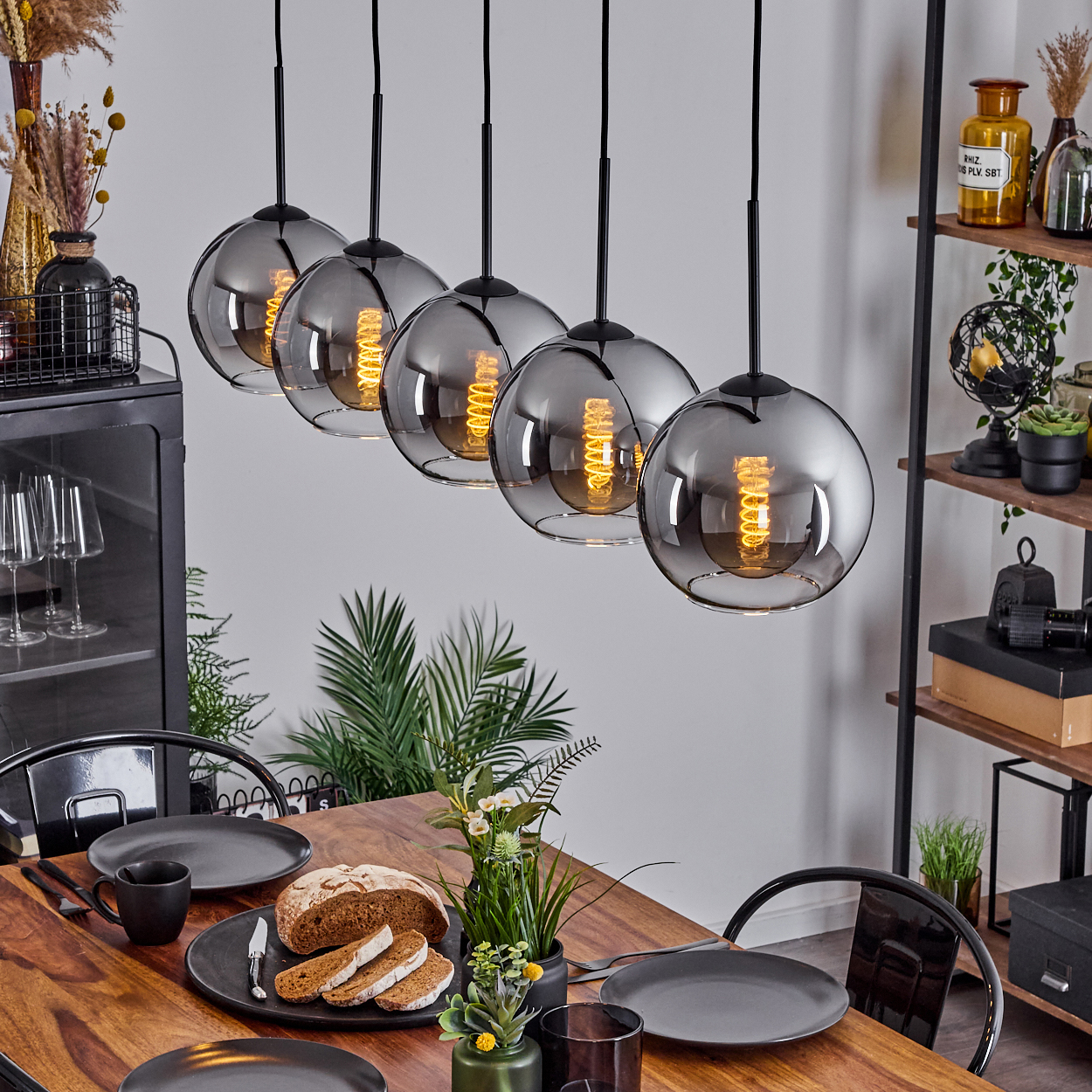 Esstisch-Set mit Pendelleuchten darüber. Teller, Essen und Dekorationen auf dem Tisch. Schwarze Stühle im Hintergrund.