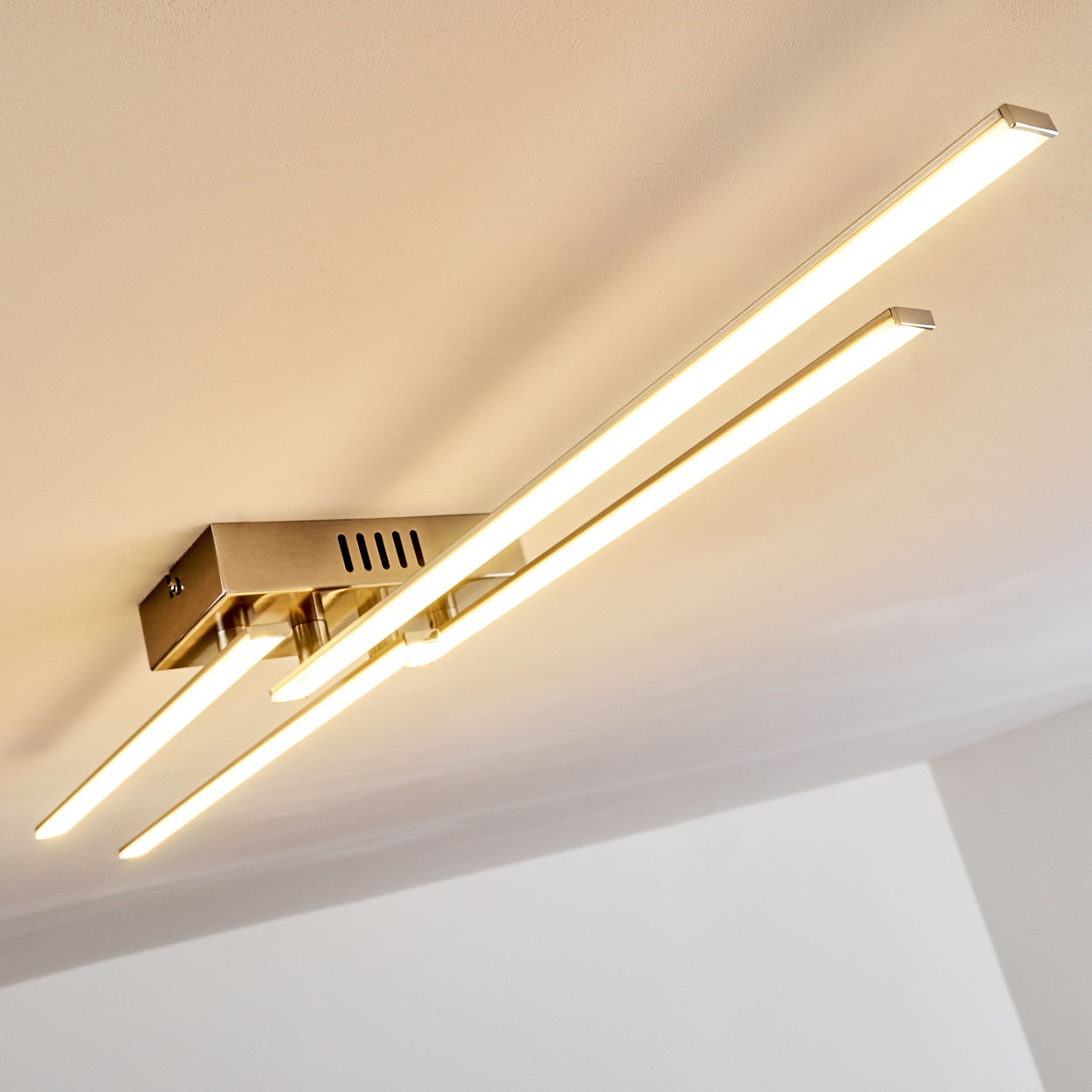 Deckenleuchte mit drei rechteckigen Lichtbalken, die warmes Licht aussenden. Die Leuchte ist metallisch, die Decke ist weiß.