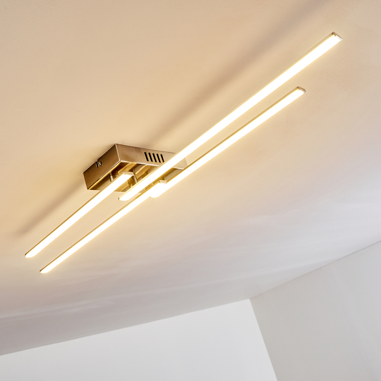 Deckenleuchte mit drei rechteckigen Lichtbalken, die warmes Licht aussenden. Die Leuchte ist metallisch, die Decke ist weiß.