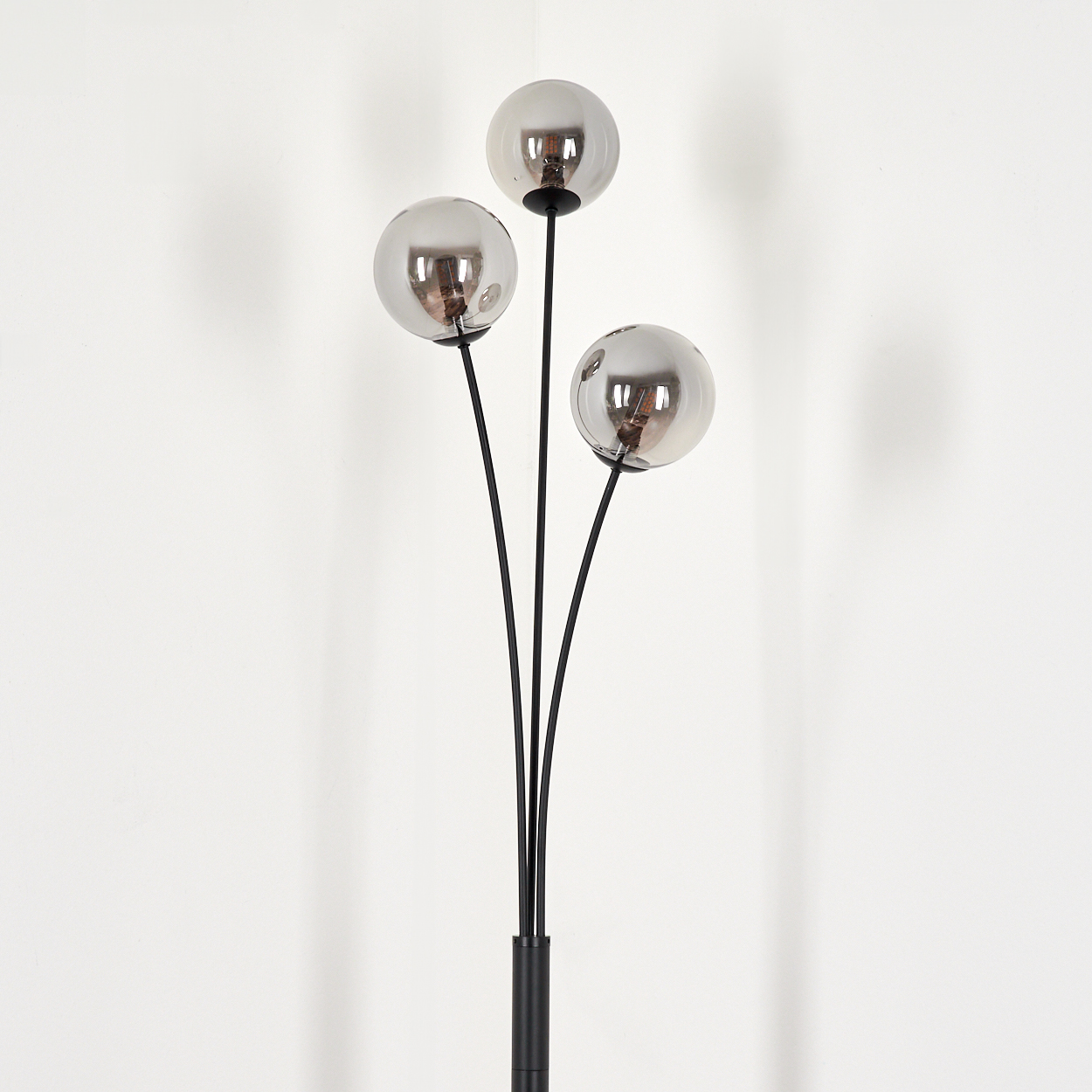 Eine Stehlampe mit drei silbernen Kugeln auf schwarzen Stielen vor einer weißen Wand. Klare Glühbirnen sind sichtbar.