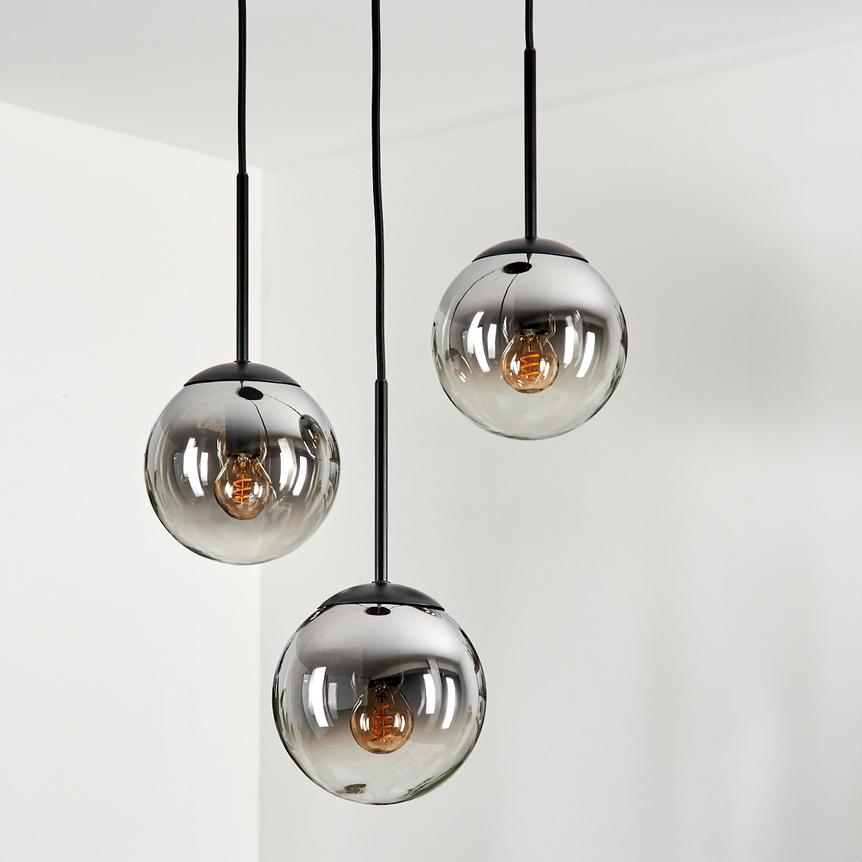 Drei Pendelleuchten mit Glaskugeln hängen vor einer weißen Wand, mit warmen Glühbirnen.