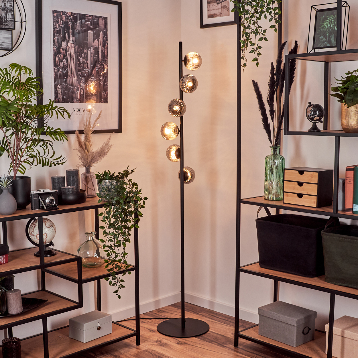 Eine schwarze Stehlampe mit Glaskugelleuchten steht in einer Ecke. Regale und Kunstwerke sind im Hintergrund.