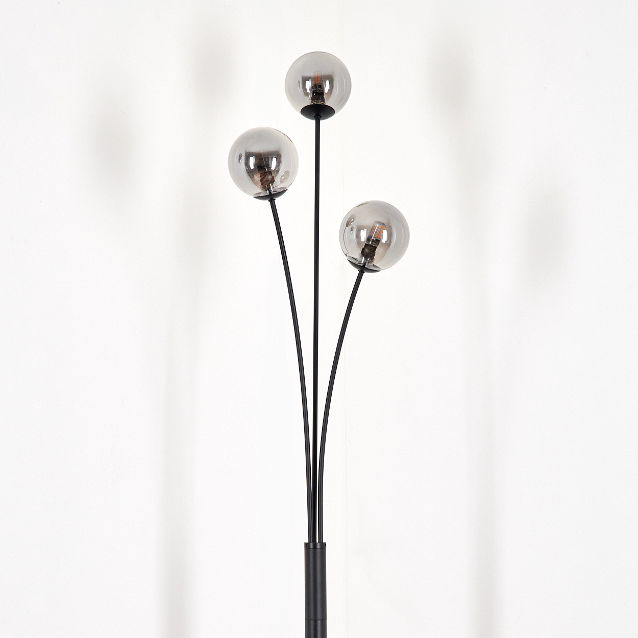 Eine Stehlampe mit drei grauen Kugeln und einem schwarzen Stiel, vor einer weißen Wand.