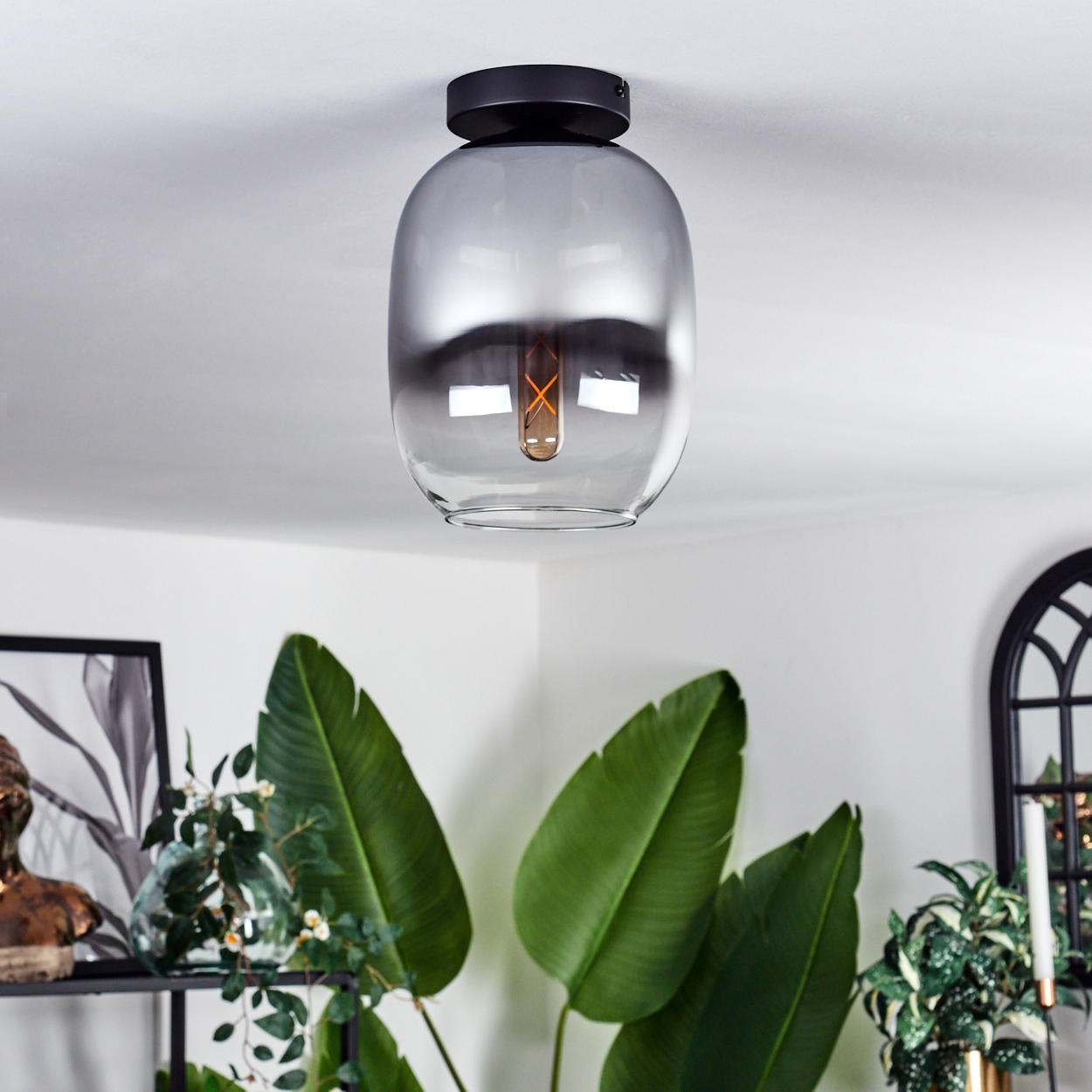 Eine Glaslampe mit einer Glühbirne im Inneren, die von der Decke hängt, mit grünen Pflanzen im Vordergrund.