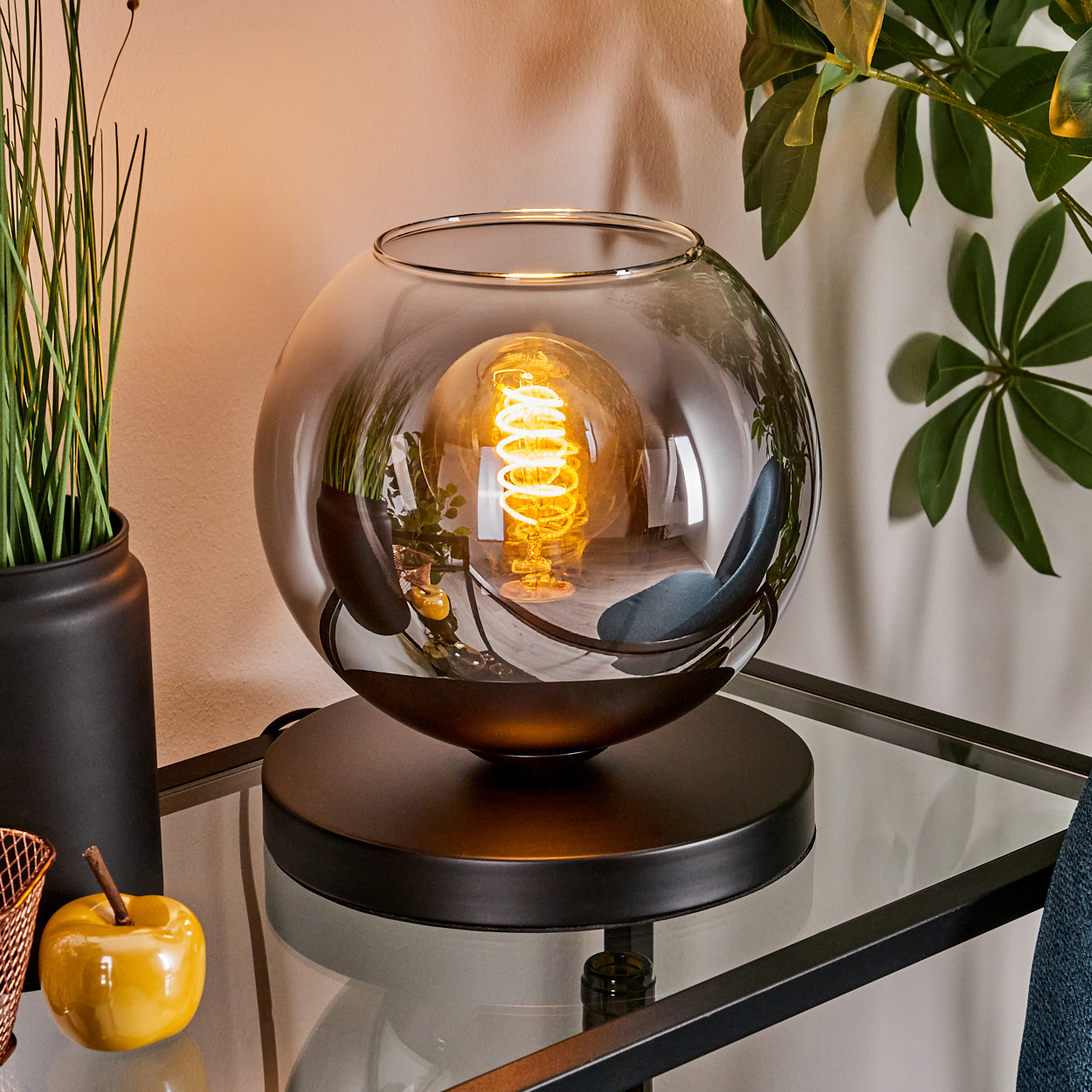 Eine Tischlampe mit einer Glühbirne in einer Glaskugel, die auf einem Tisch steht. Ein gelber Apfel liegt auf dem Tisch.