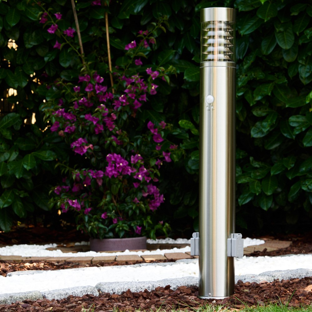 Eine Außenleuchte aus Edelstahl. Sie steht in einem Garten mit Büschen und weißem Kies. Sie hat einen Sensor.