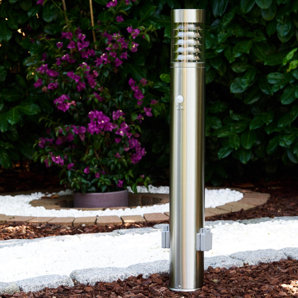 Eine Außenleuchte aus Edelstahl mit Sensor. Sie steht in einem Garten mit Büschen und weißem Kies.