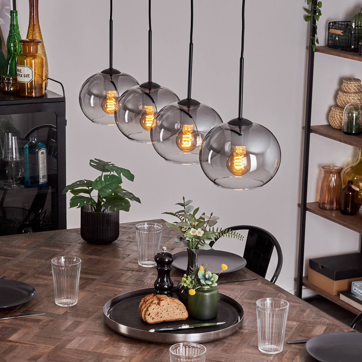 Vier Pendelleuchten mit rauchfarbenen Glaskugeln. Sie hängen über einem Holztisch mit Gläsern und Essen.