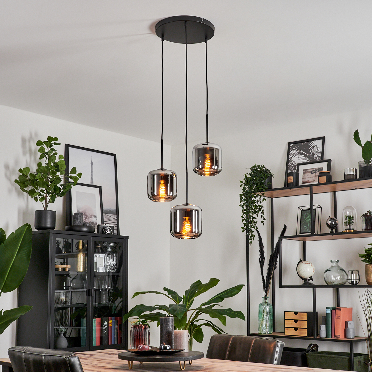 Drei Pendelleuchten mit Glasschirmen hängen über einem Tisch. Pflanzen und Dekorationen sind im Hintergrund.