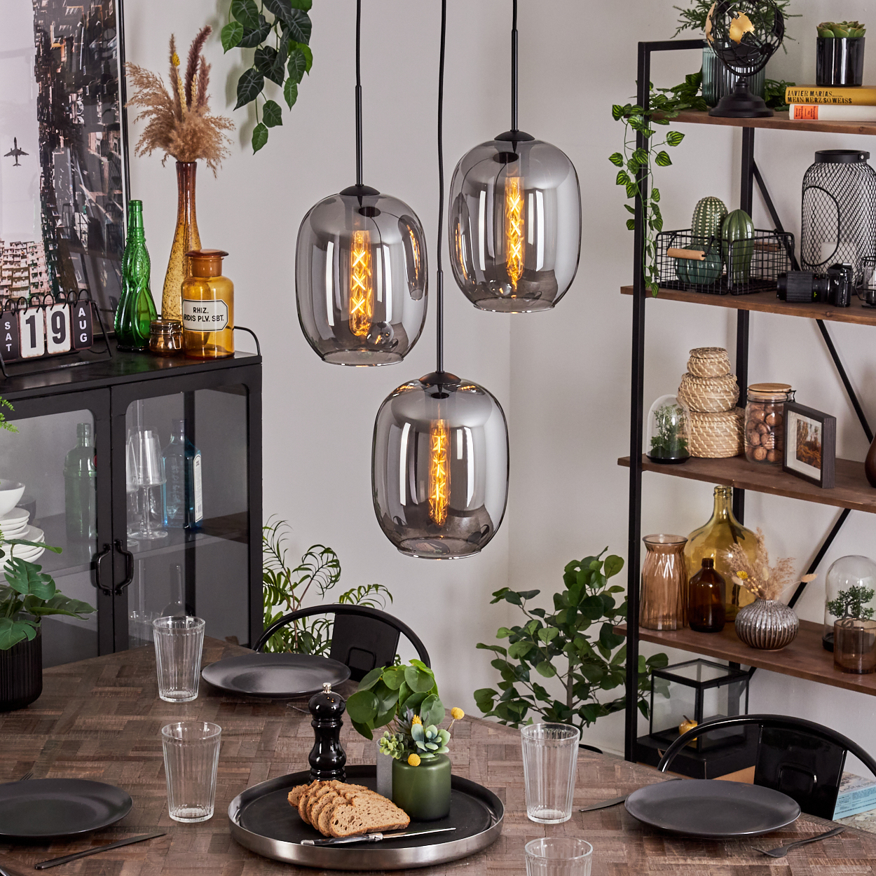 Esszimmer mit drei Pendelleuchten, Glasschirmen, Pflanzen und Dekoration, gedeckt für das Abendessen.