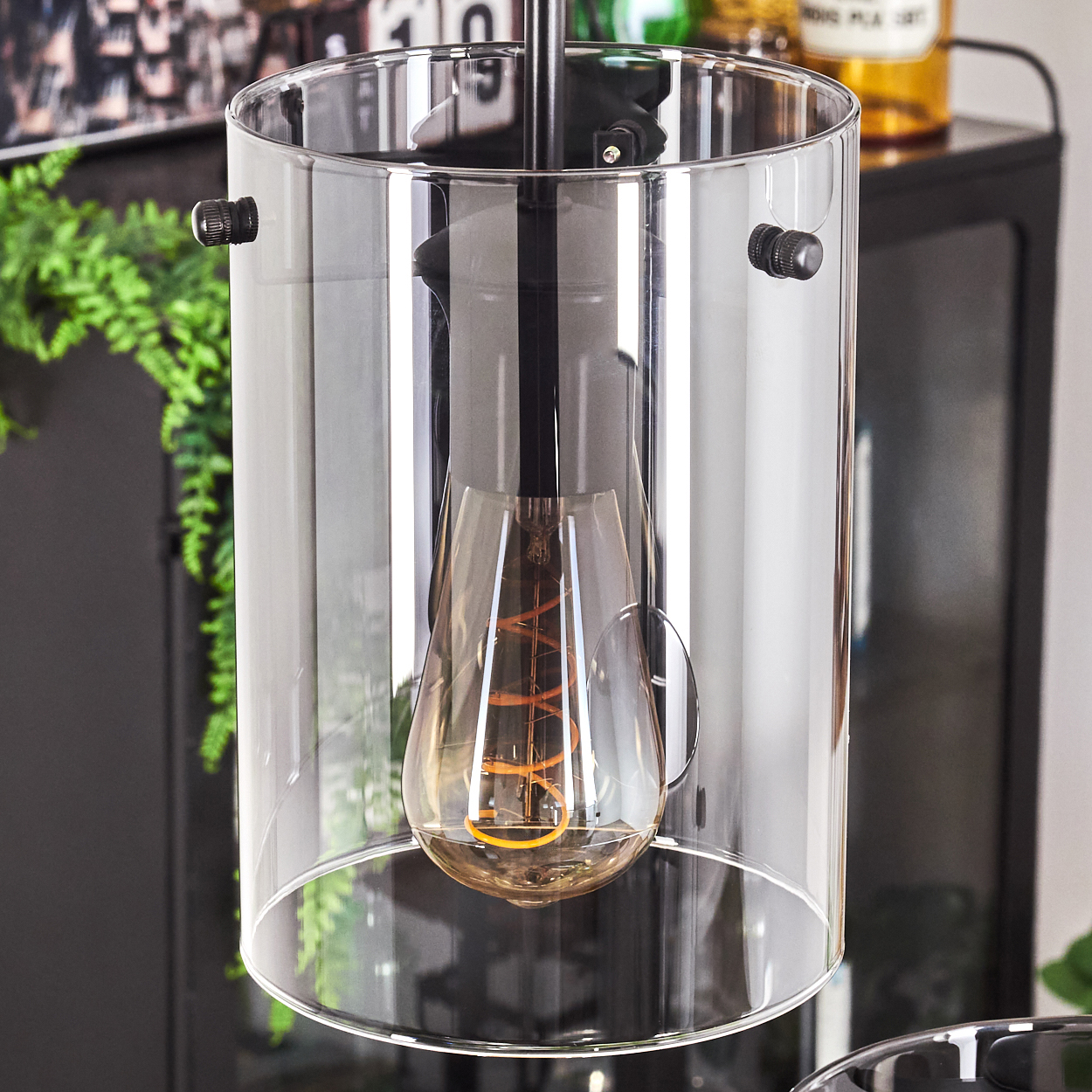 Nahaufnahme einer zylindrischen Lampe mit transparentem Glasschirm und sichtbarer Glühbirne. Schwarze Details sichtbar.