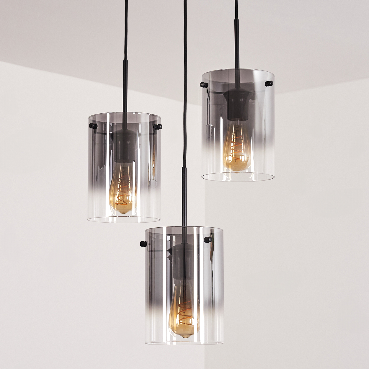 Drei zylindrische rauchfarbene Glas-Pendelleuchten mit sichtbaren Glühbirnen, angeordnet vor einer weißen Wand.