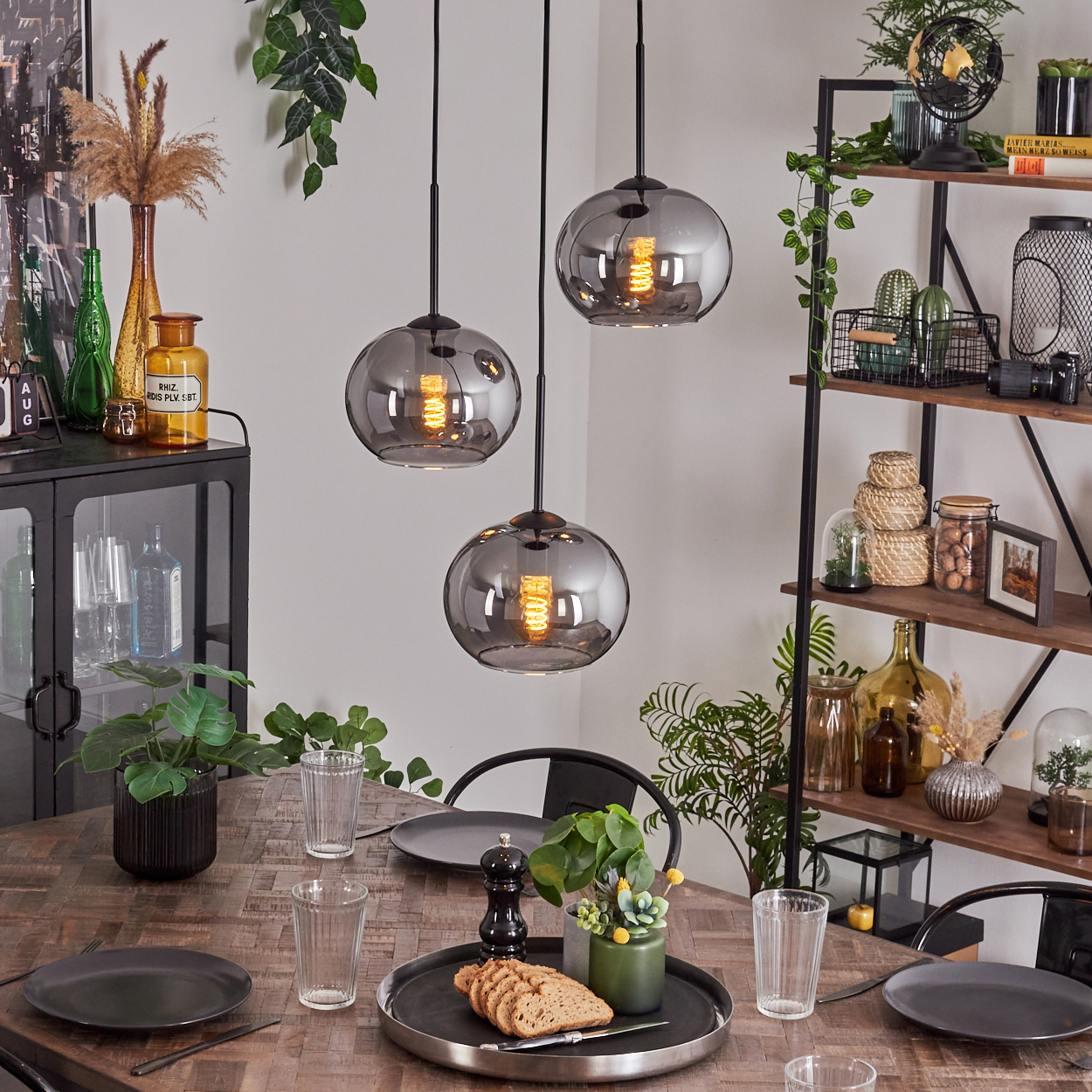 Esszimmer mit drei rauchfarbenen Glaskugel-Pendelleuchten über einem Holztisch, Pflanzen und Dekoration.