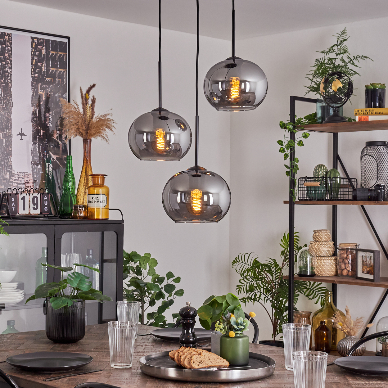 Esszimmer mit Holztisch, drei Pendelleuchten und verschiedenen Dekorationsartikeln auf Regalen.