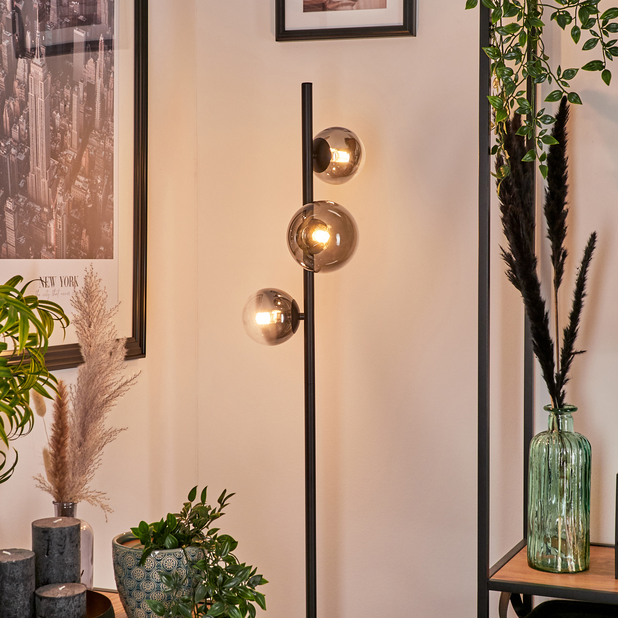 Eine Stehlampe mit drei Kugelleuchten, mit Dekor und Pflanzen. Die Lichter sind an, und die Stange ist schwarz.