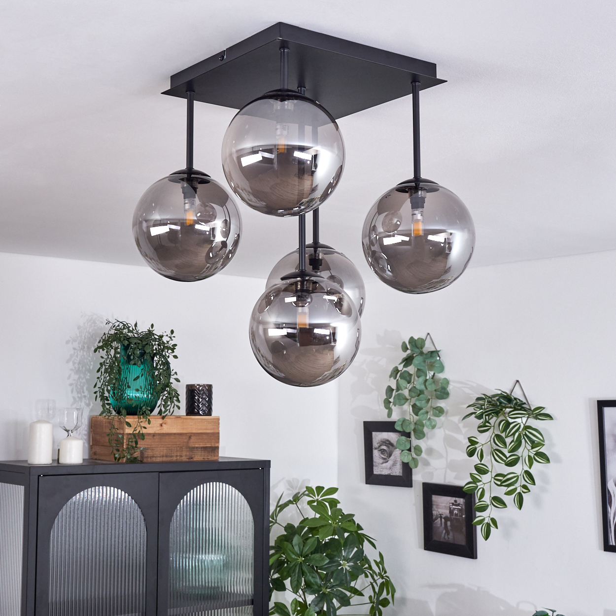 Deckenlampe mit fünf Glaskugeln. Pflanzen und Möbel im Hintergrund. Das Licht beleuchtet den Raum.