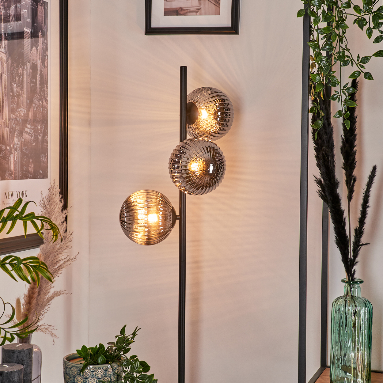 Eine Stehlampe mit drei verchromten Kugeln mit strukturiertem Glas. Schwarze Stange. Innenraum mit Pflanzen und Dekor.