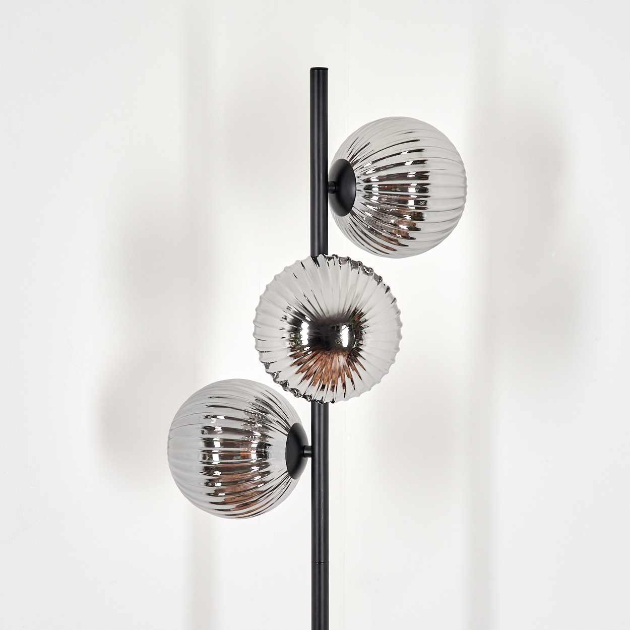 Eine moderne Stehlampe mit drei silbernen, gerippten Glaskugeln. Ein schwarzer Pfosten trägt die Kugeln auf weißem Grund.